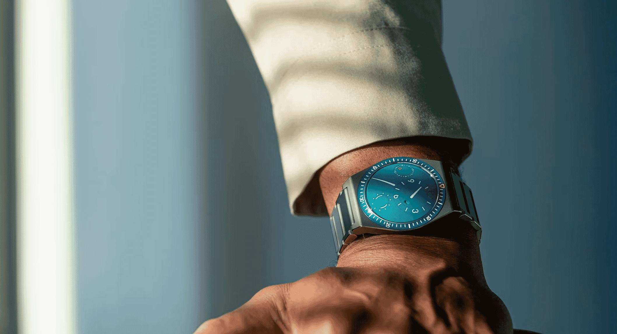 Man's wrist wearing a sleek metallic watch with a blue dial against a formal background.