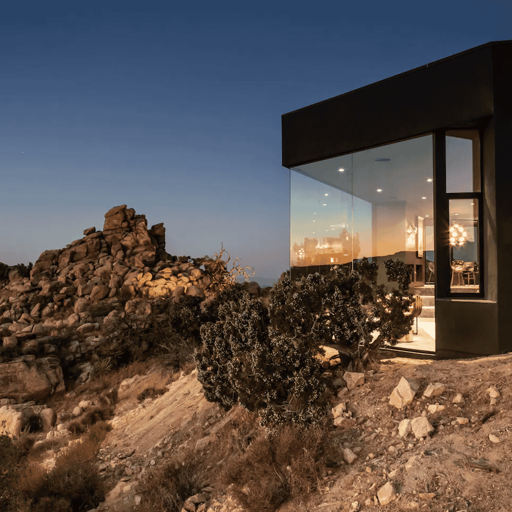 A modern glass-walled house perched on a rocky desert hillside at twilight with glowing interiors.