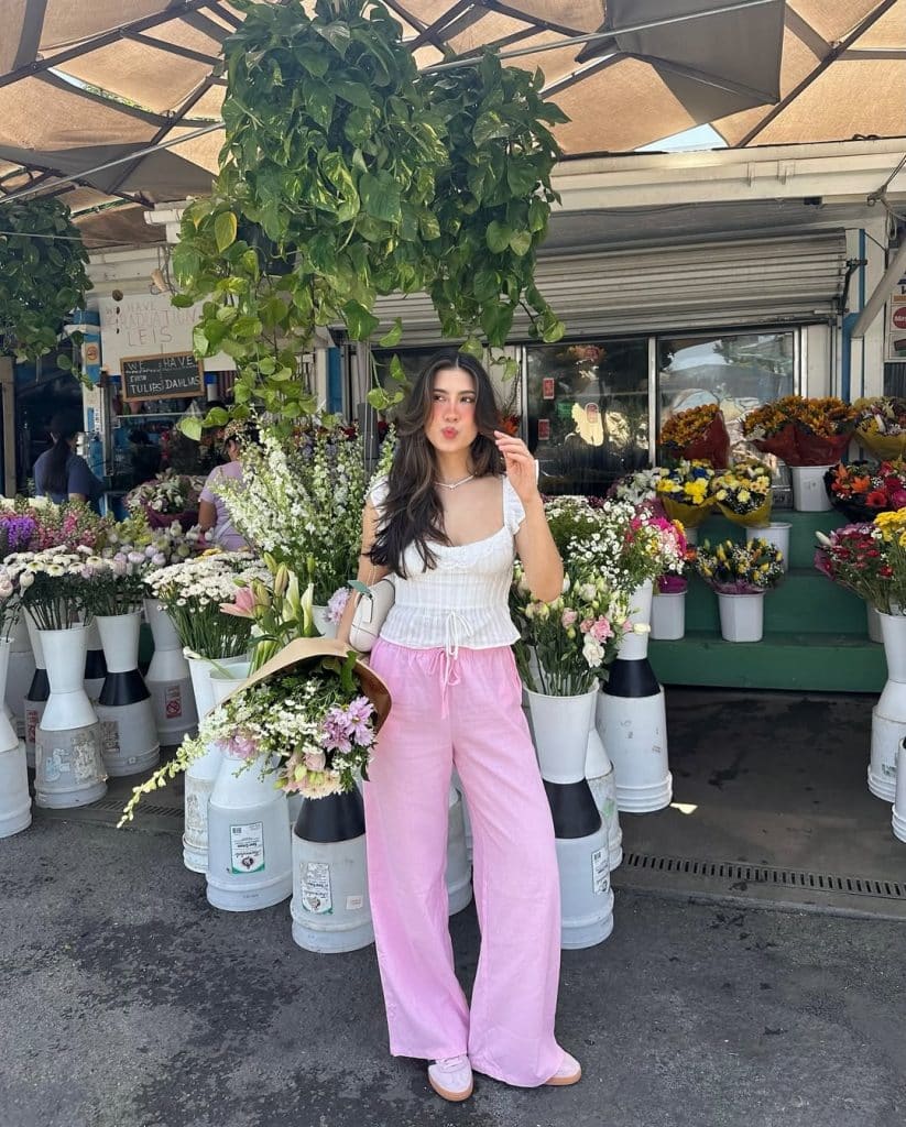 A woman in pink wide-leg pants and a white ruffled top posing with flowers at an outdoor market.