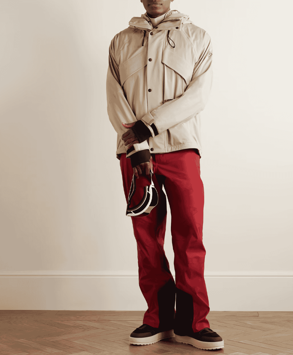A model in a beige hooded jacket and vibrant red ski pants, holding goggles against a minimalist backdrop.