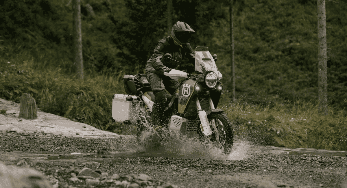 An adventure motorcycle splashing through a muddy trail in a forest.
