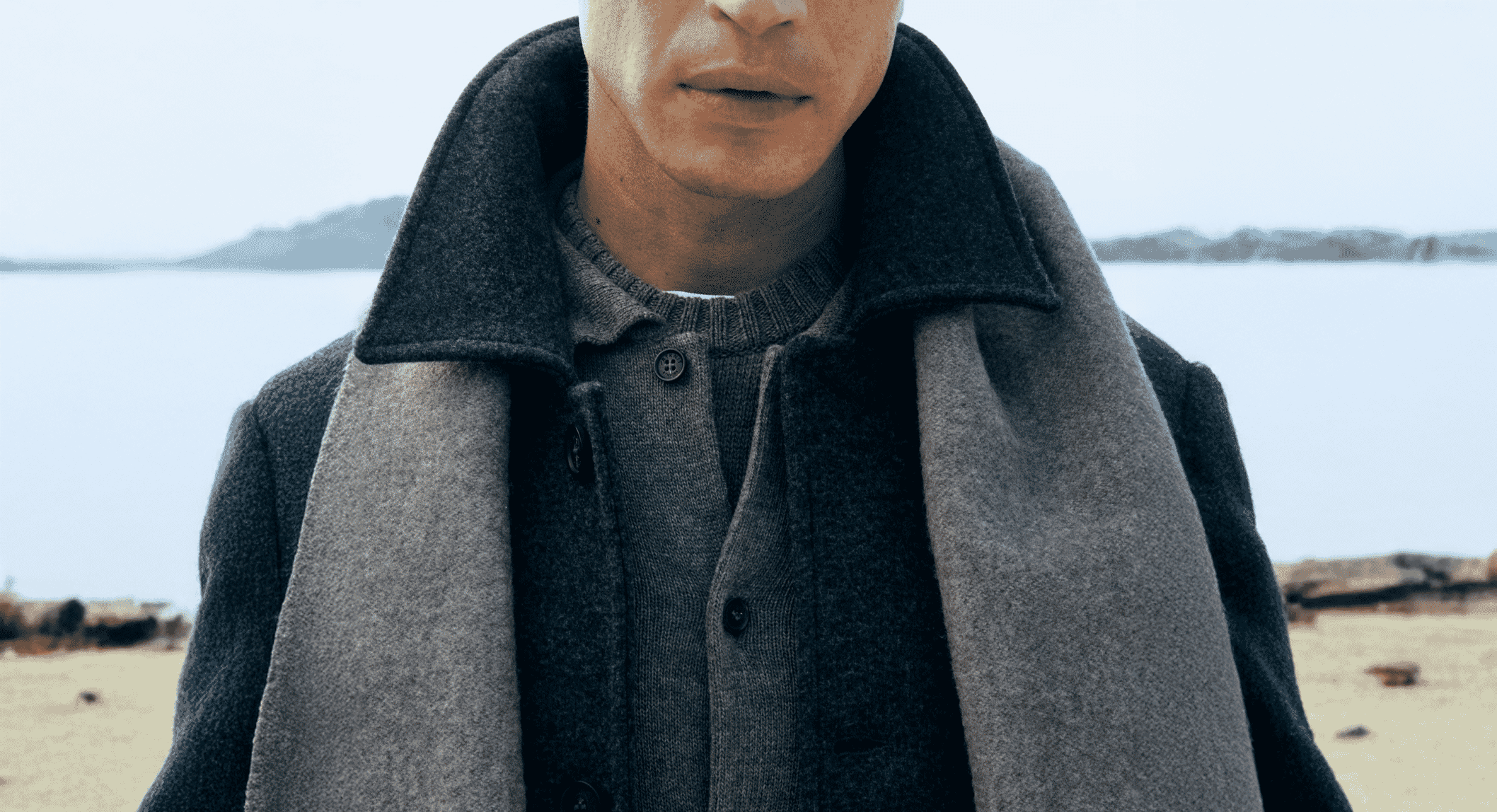 A man in a dark layered wool coat and knit sweater at a beach shoreline.