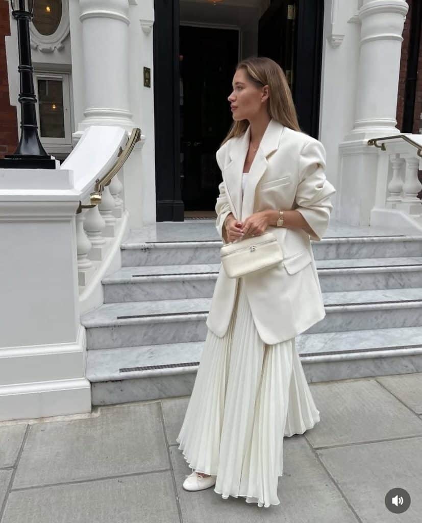 A woman in a structured white blazer and pleated white skirt standing on marble steps.