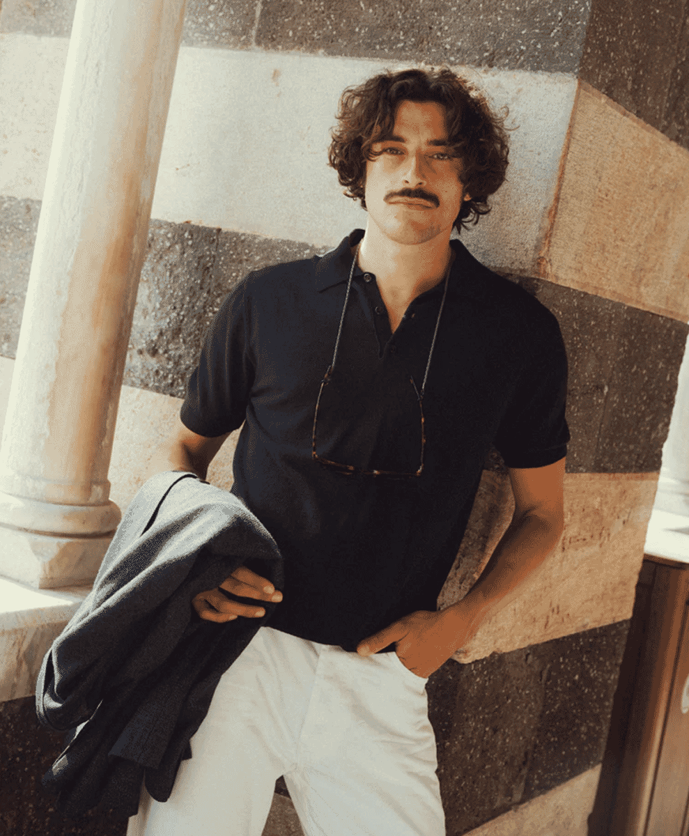 A man in a navy polo shirt and white trousers leaning against a stone column.