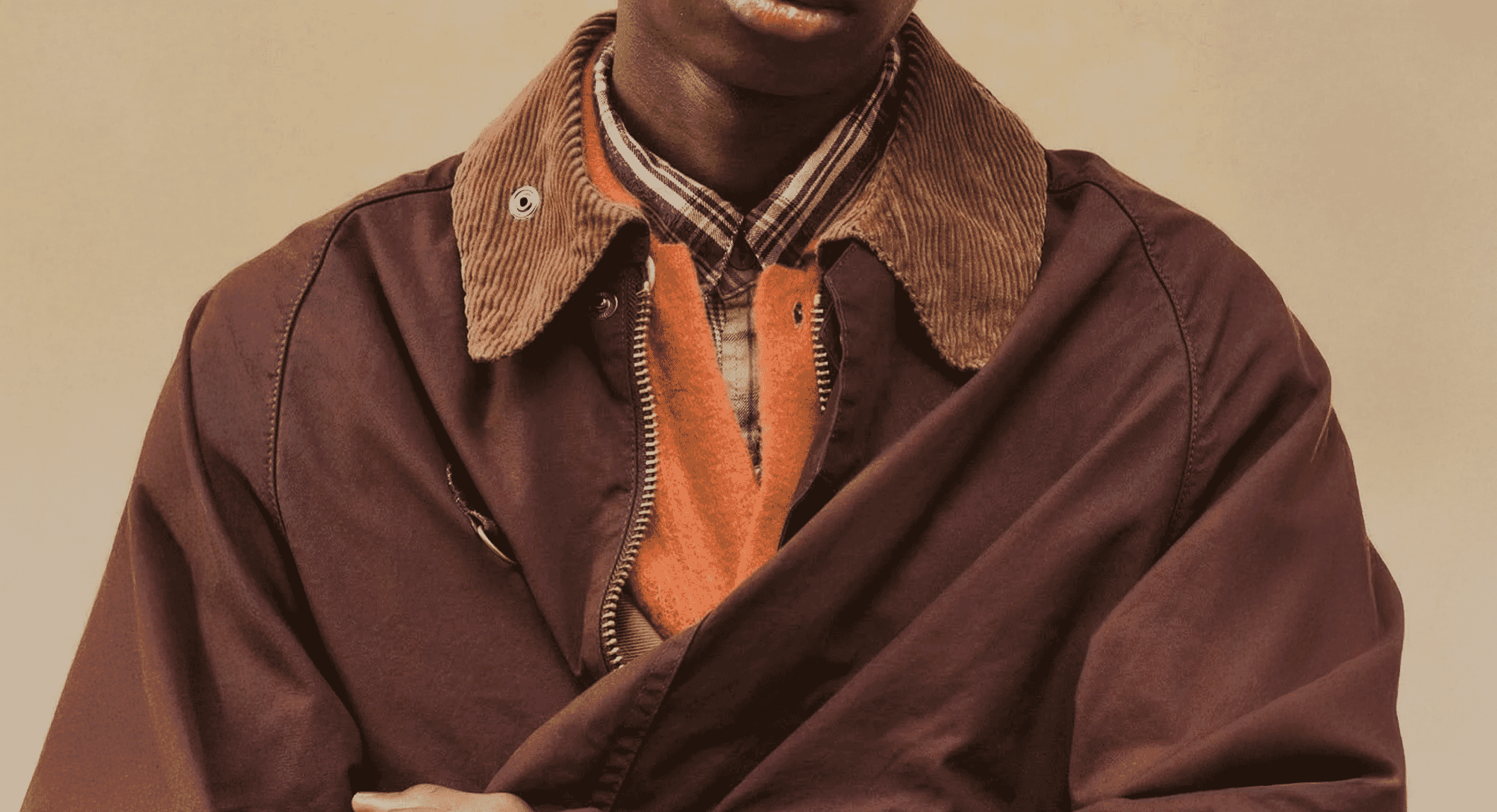 A man wearing a layered outfit featuring a brown jacket, orange sweater, and plaid shirt.