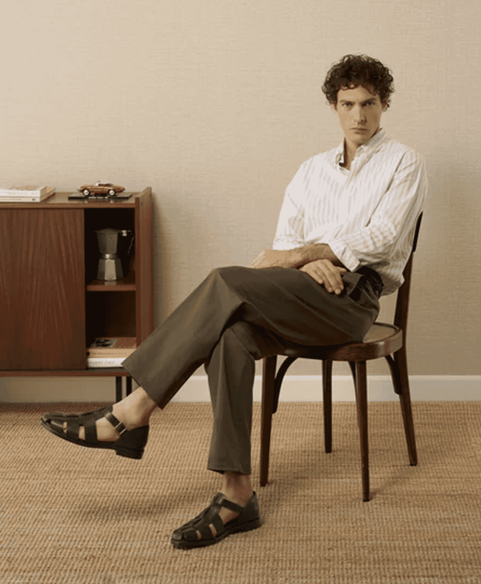 A man sitting on a chair wearing black leather sandals with wide-leg trousers.