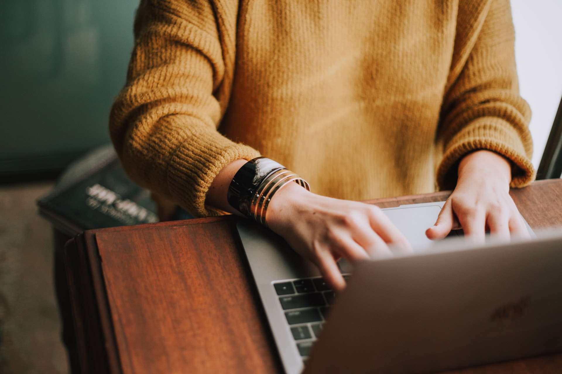 A person in a cozy mustard-yellow ribbed sweater with layered bracelets types on a laptop at a wooden desk.