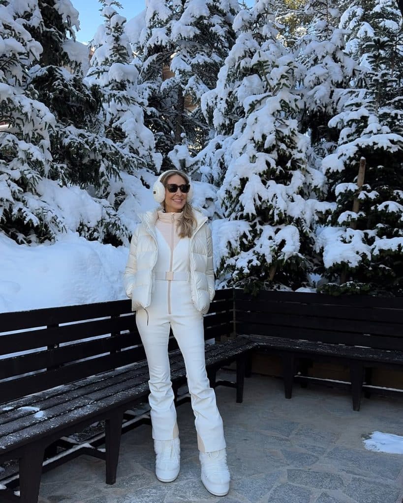 Woman in a white winter outfit and earmuffs standing by snow-covered trees.