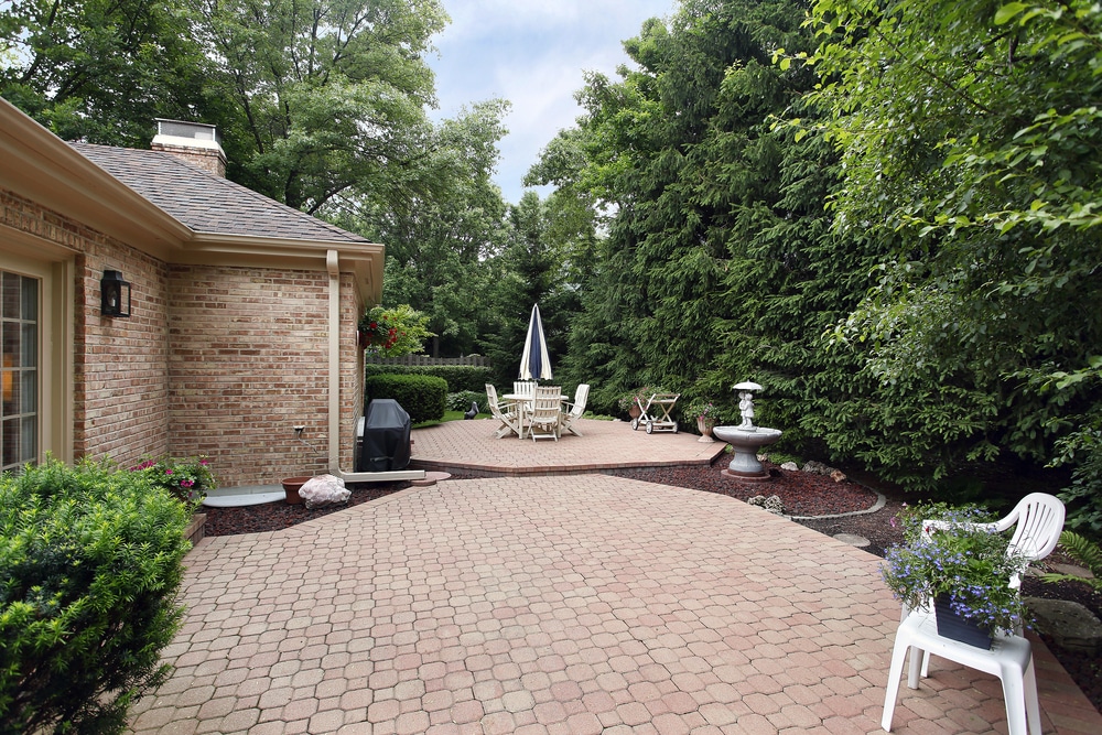 A serene brick patio with a white fountain and classic white outdoor furniture surrounded by dense plants.