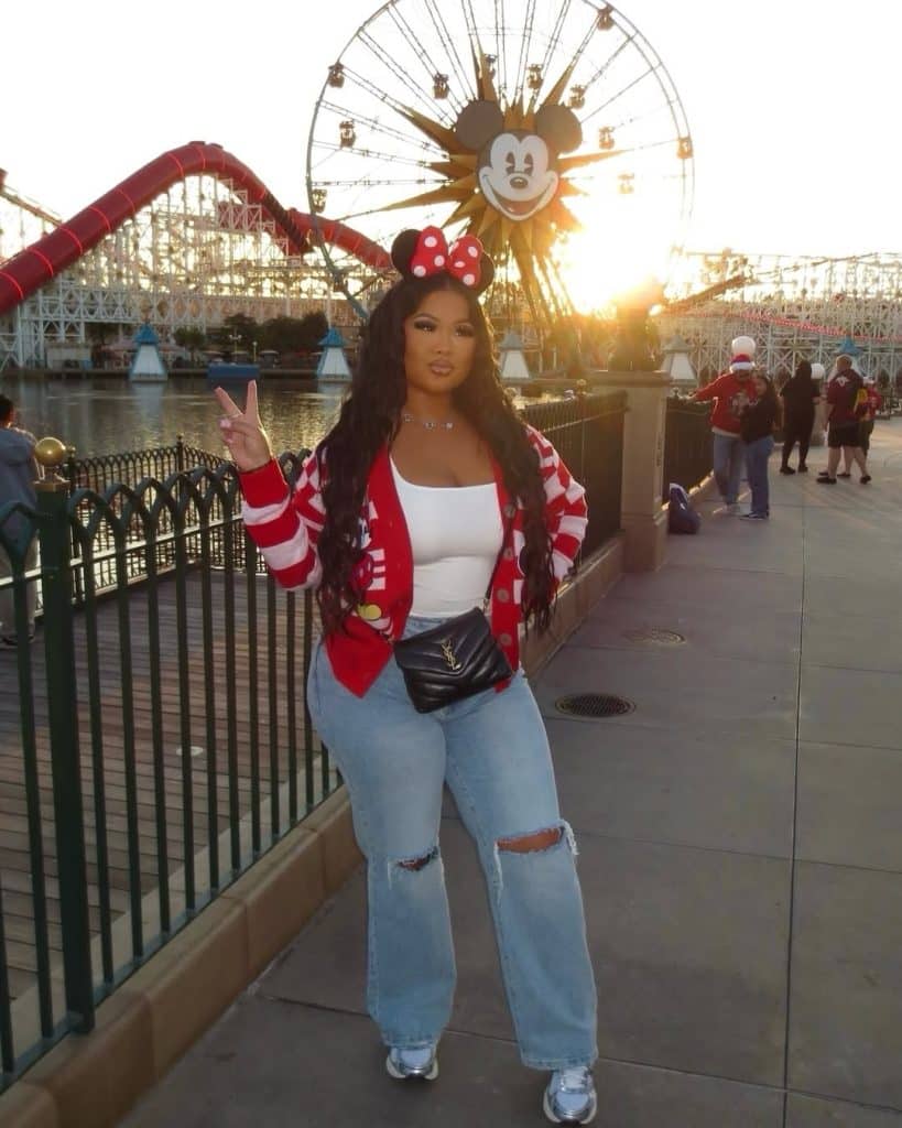 A woman in a red-and-white striped Mickey cardigan and ripped jeans at sunset with a Ferris wheel behind her.
