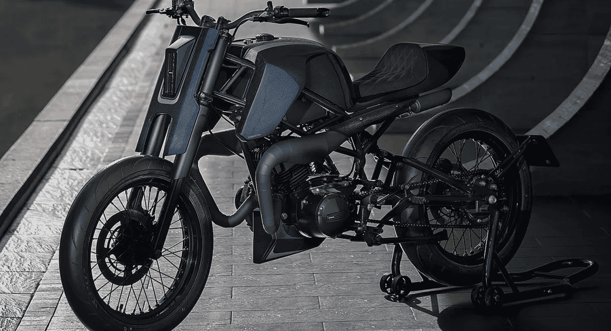 Profile view of a custom motorcycle showing the trellis frame against a curved architectural wall.