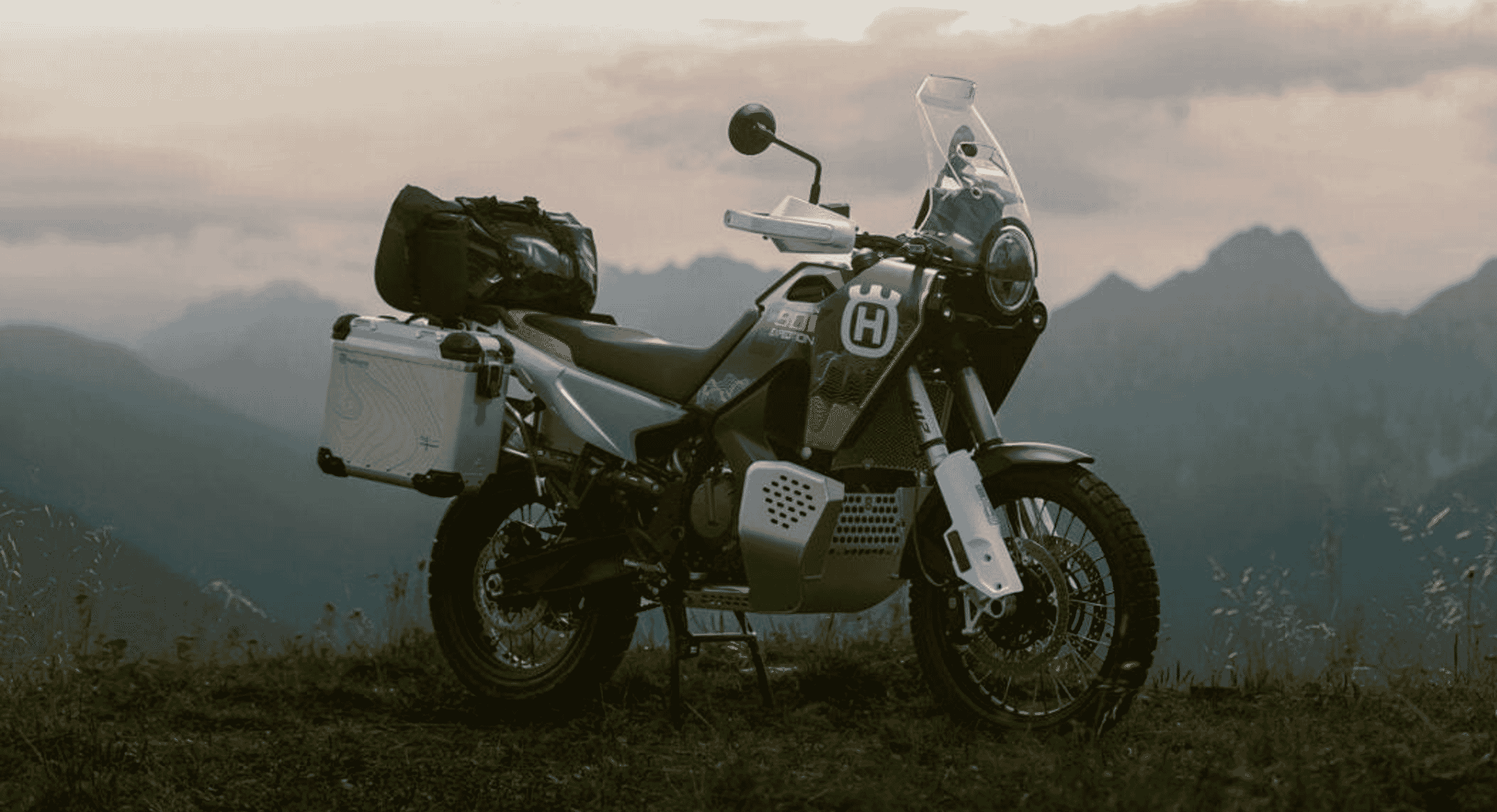 Husqvarna adventure bike equipped with luggage on a grassy mountain ridge at dusk.