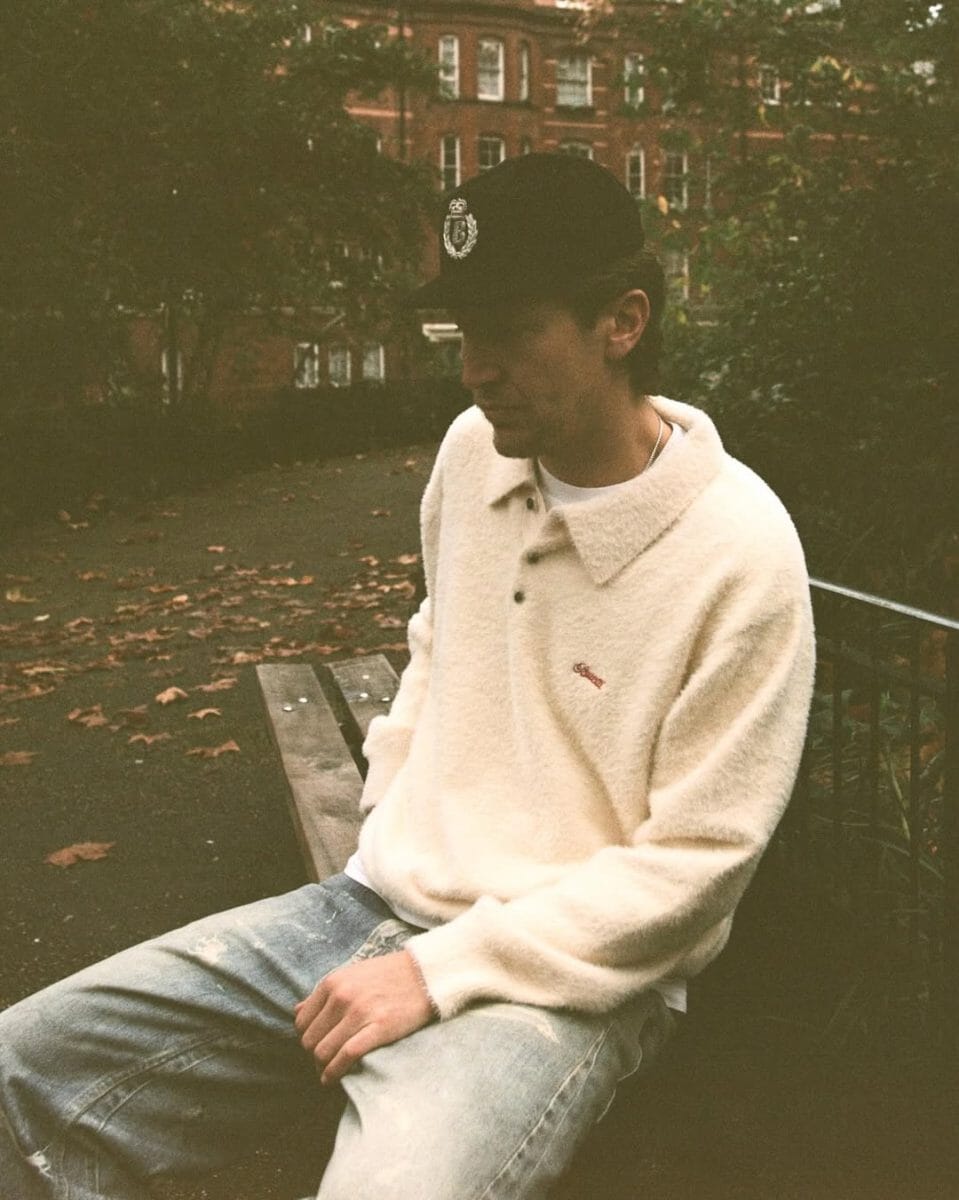 A man wearing a cream fuzzy polo sweater and a black cap sitting on a bench.