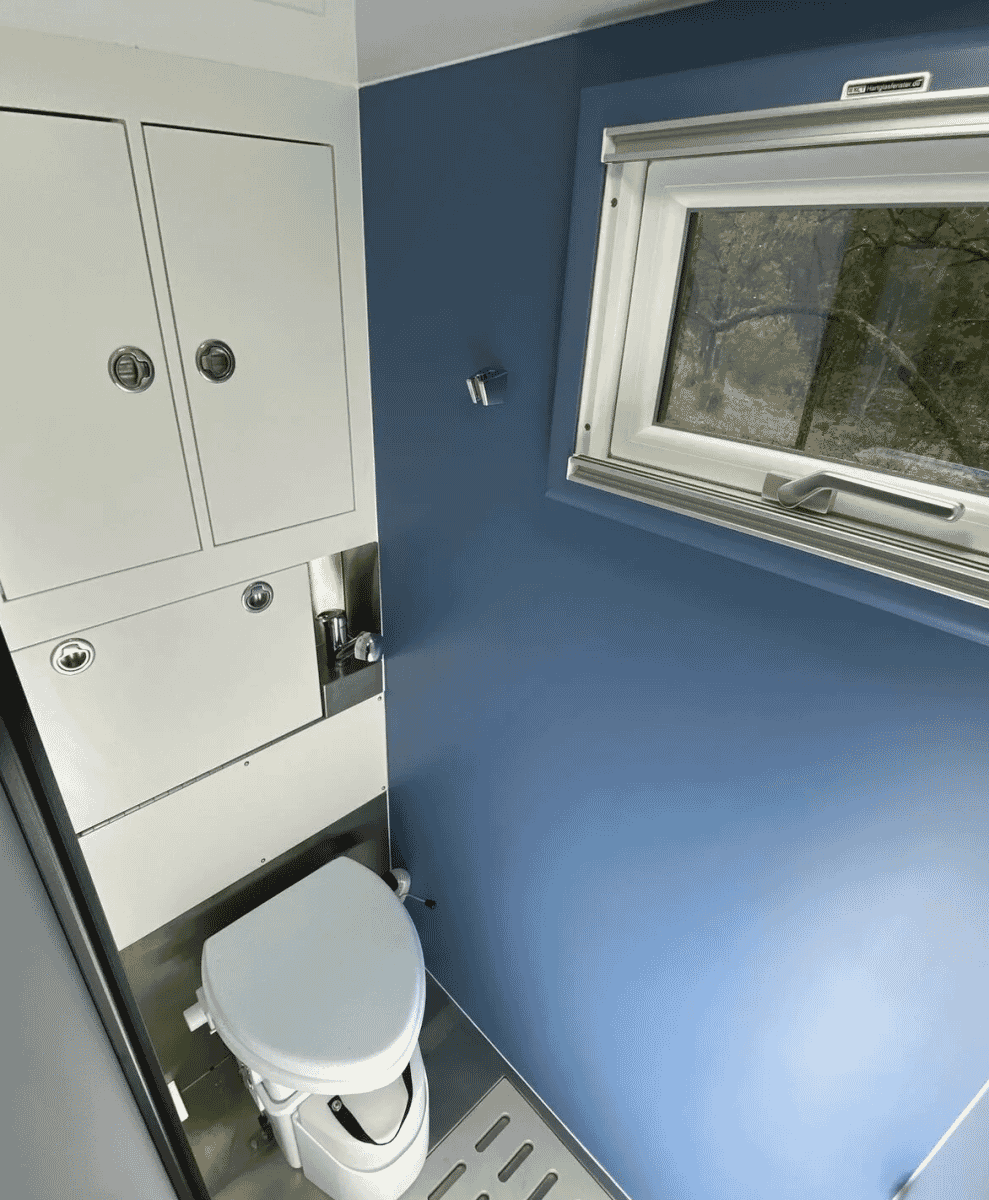 A compact blue bathroom inside the RV featuring modern fixtures and a window with a forest view.