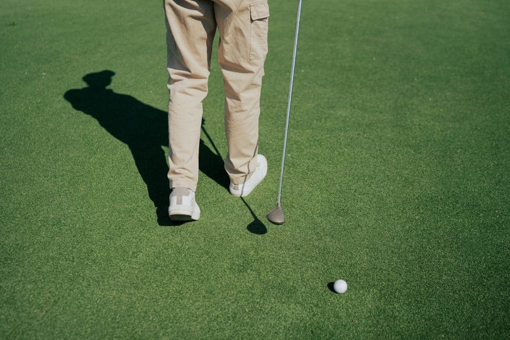 Golfer in beige cargo-style pants holding a putter on a sunlit putting green.
