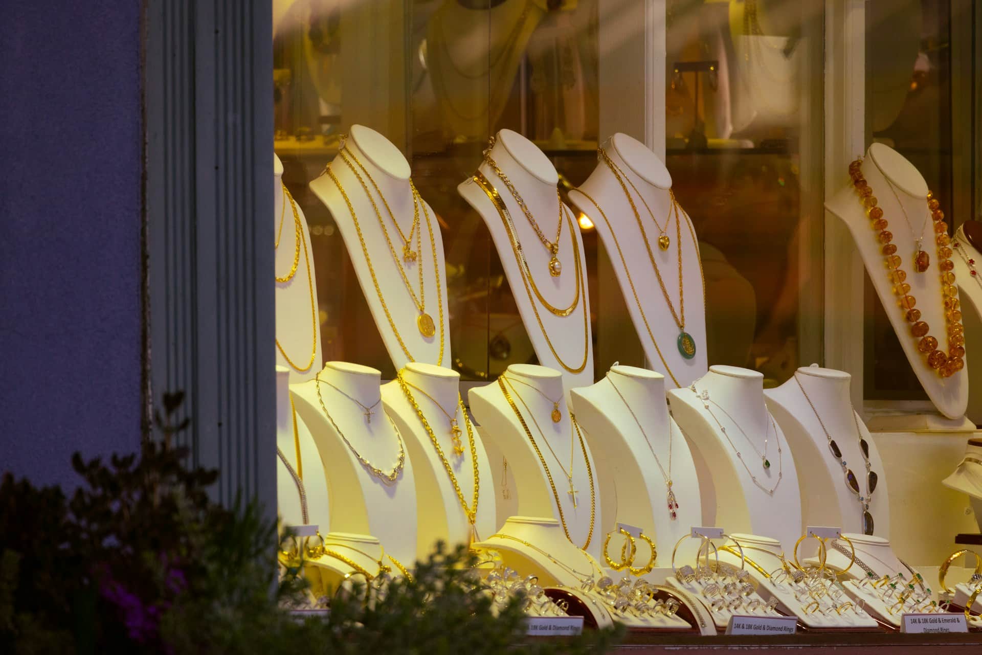 A jewelry storefront window display featuring gold and silver necklaces on white busts with warm lighting.