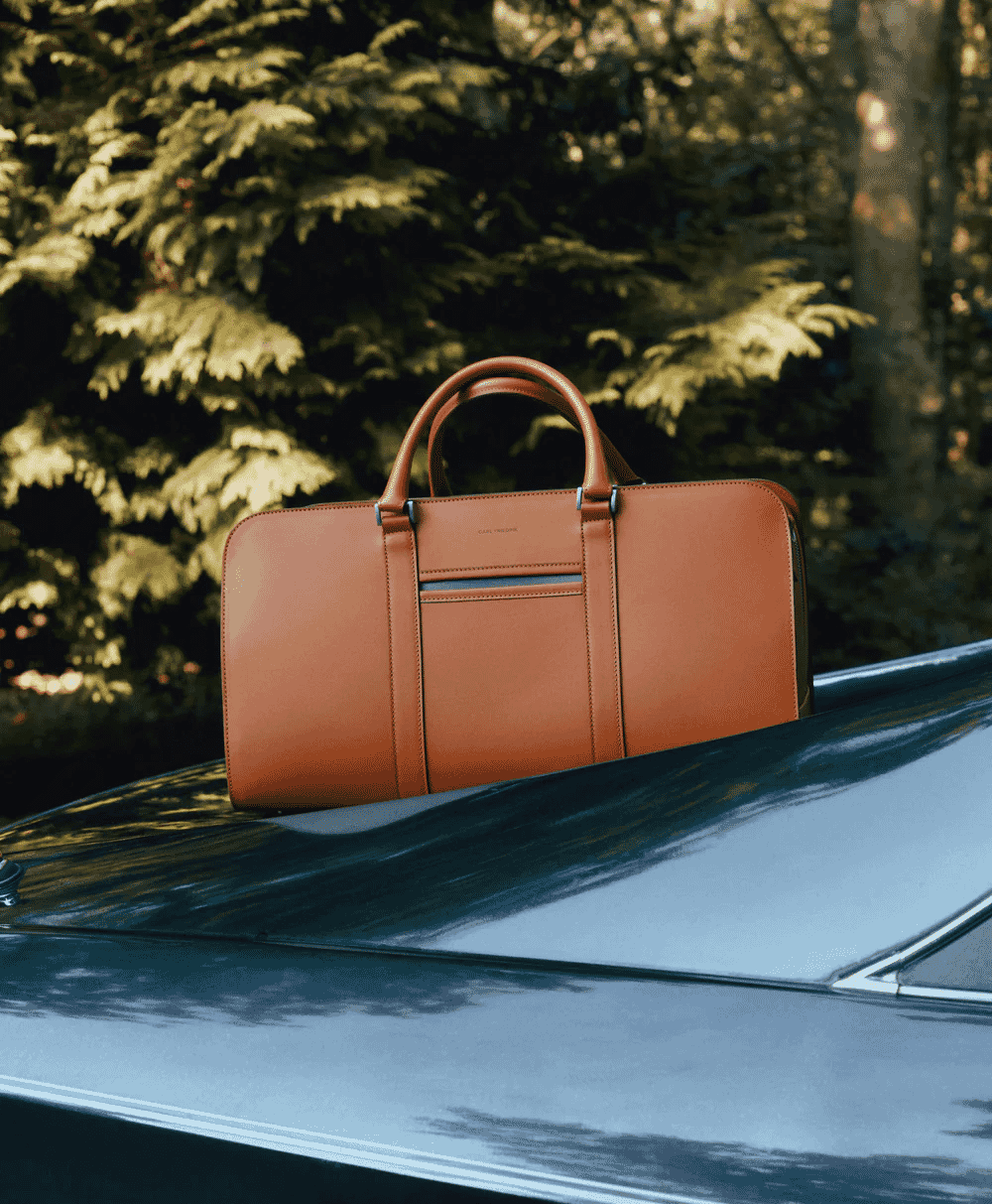 A tan leather duffel bag resting on the hood of a classic car in a forest setting.