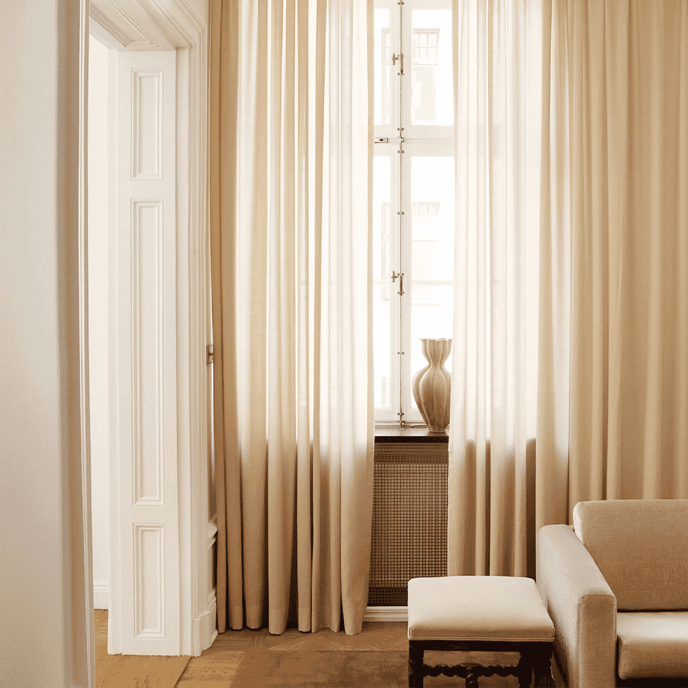 Soft cream-colored curtains framing a window next to a matching armchair and ottoman in a minimalist room.