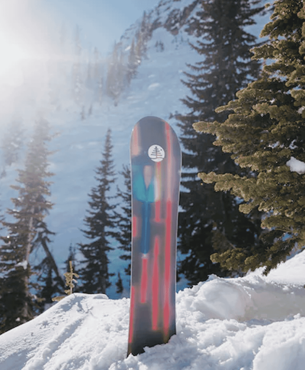 A snowboard standing upright in fresh powder snow with evergreen trees and mountain peaks in the background.
