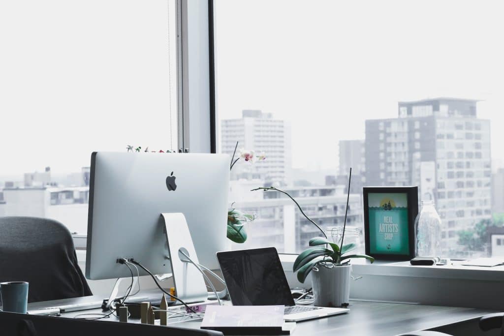A sleek, minimalist office workspace featuring an iMac and laptop near a window with an urban city view.