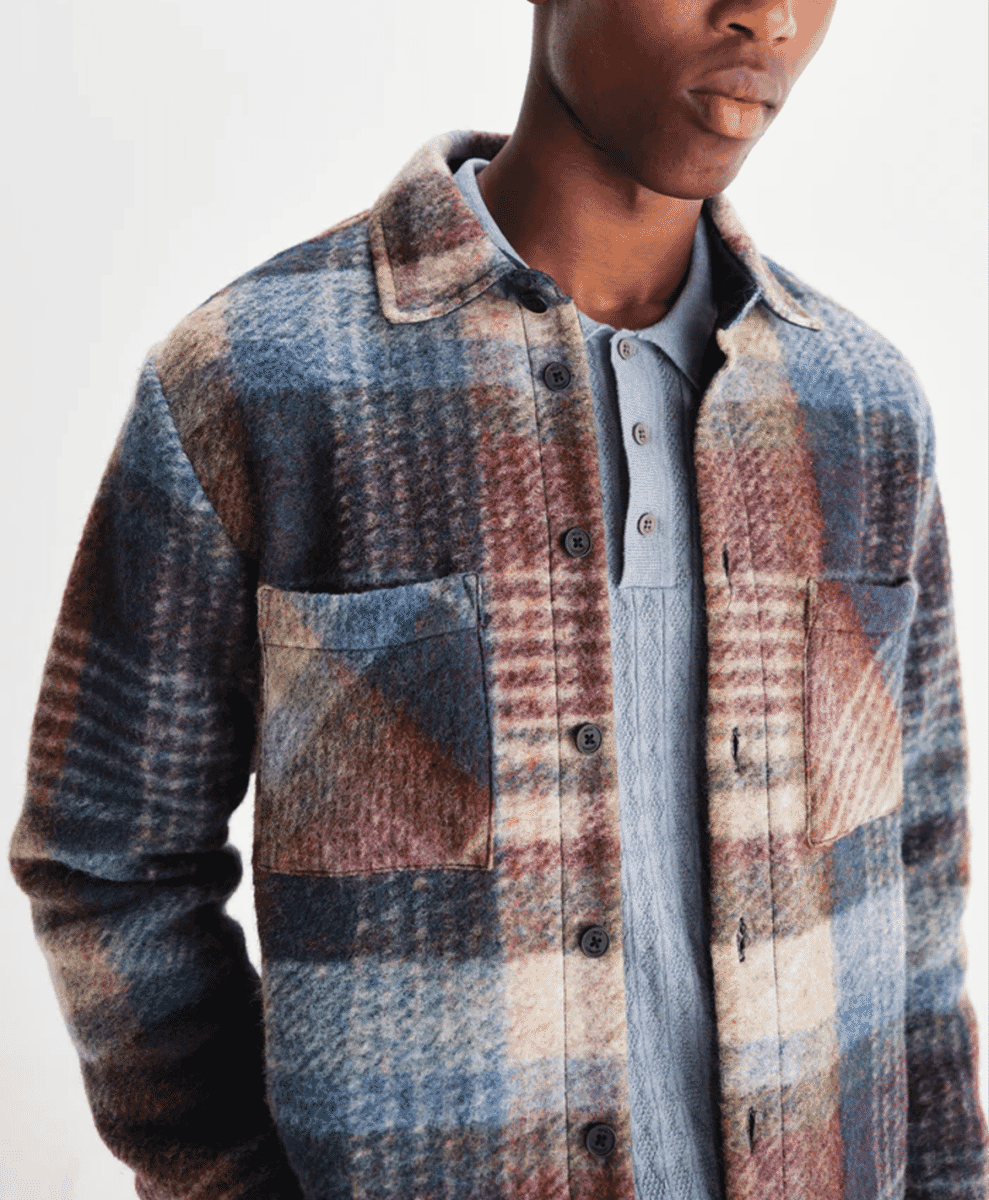 A man models a textured plaid wool shirt jacket in blue and brown tones in a studio setting.