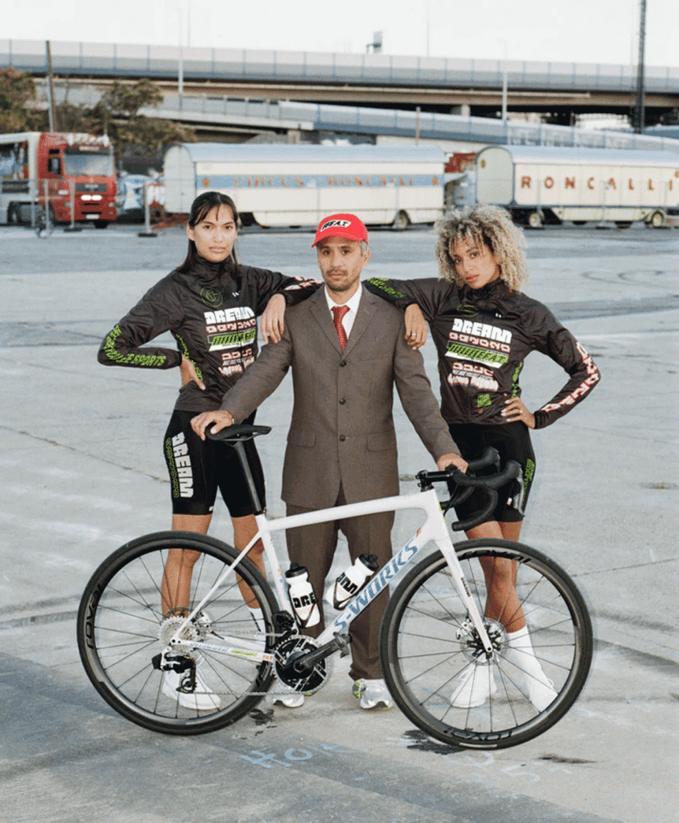 Two cyclists in black and green kits posing with a man in a brown suit and red cap alongside a white S-Works road bike.