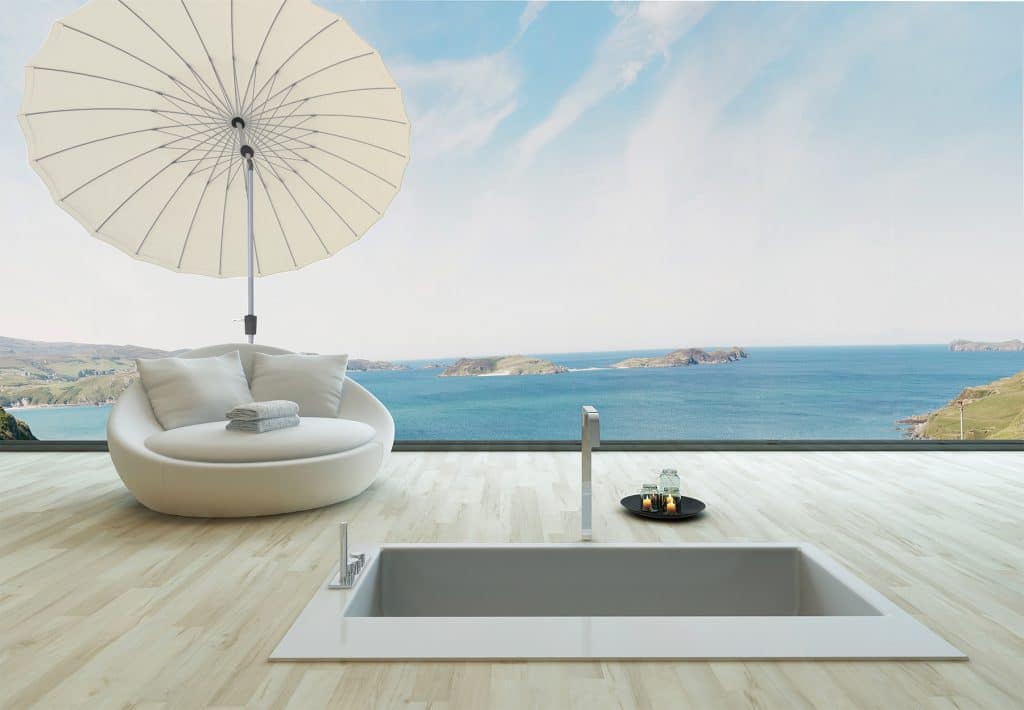 A luxury bathroom with a sunken tub and a white lounge chair under a parasol overlooking an ocean view.