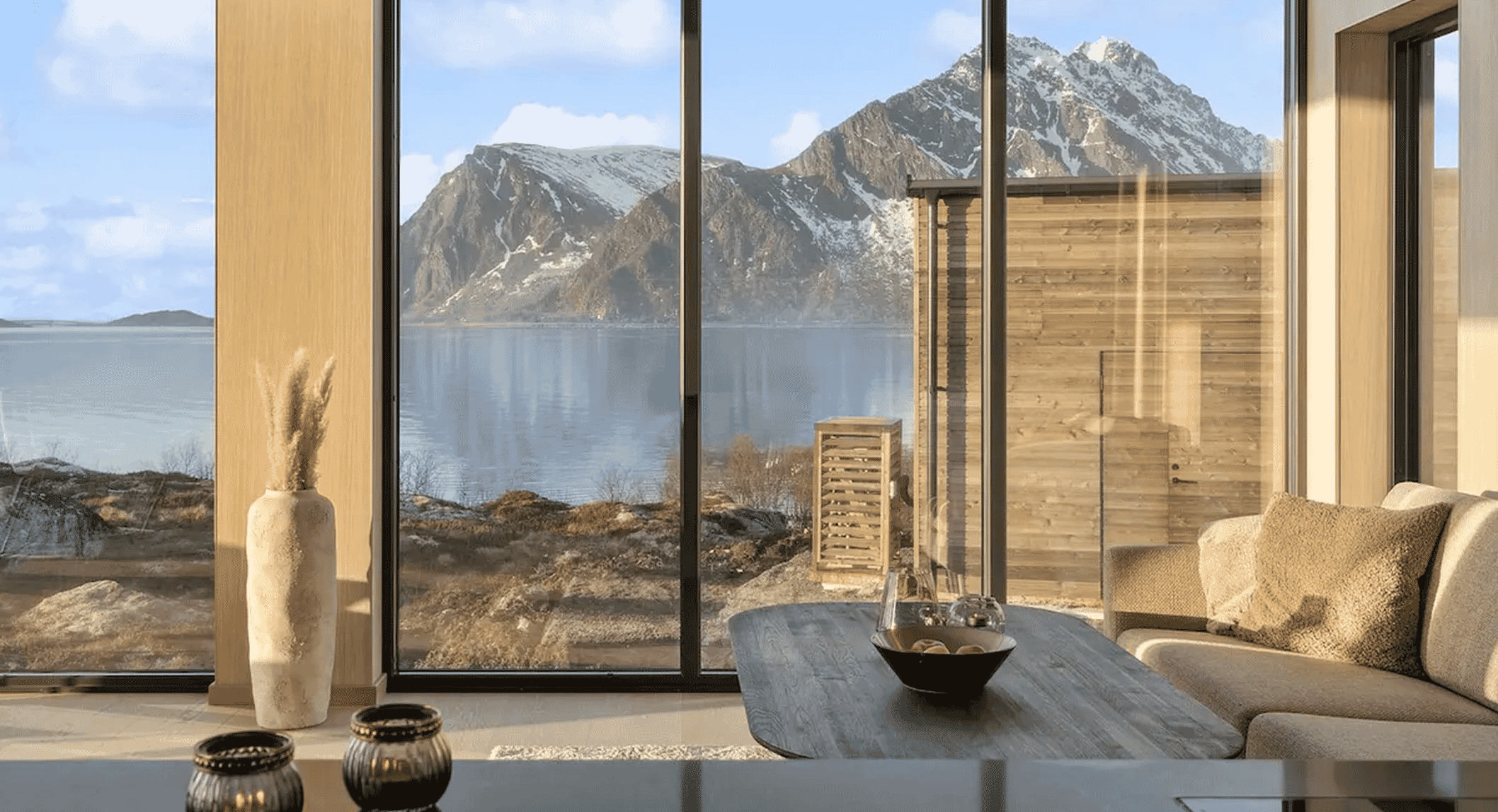 Modern living room with large glass windows overlooking snow-capped mountains and a calm fjord.