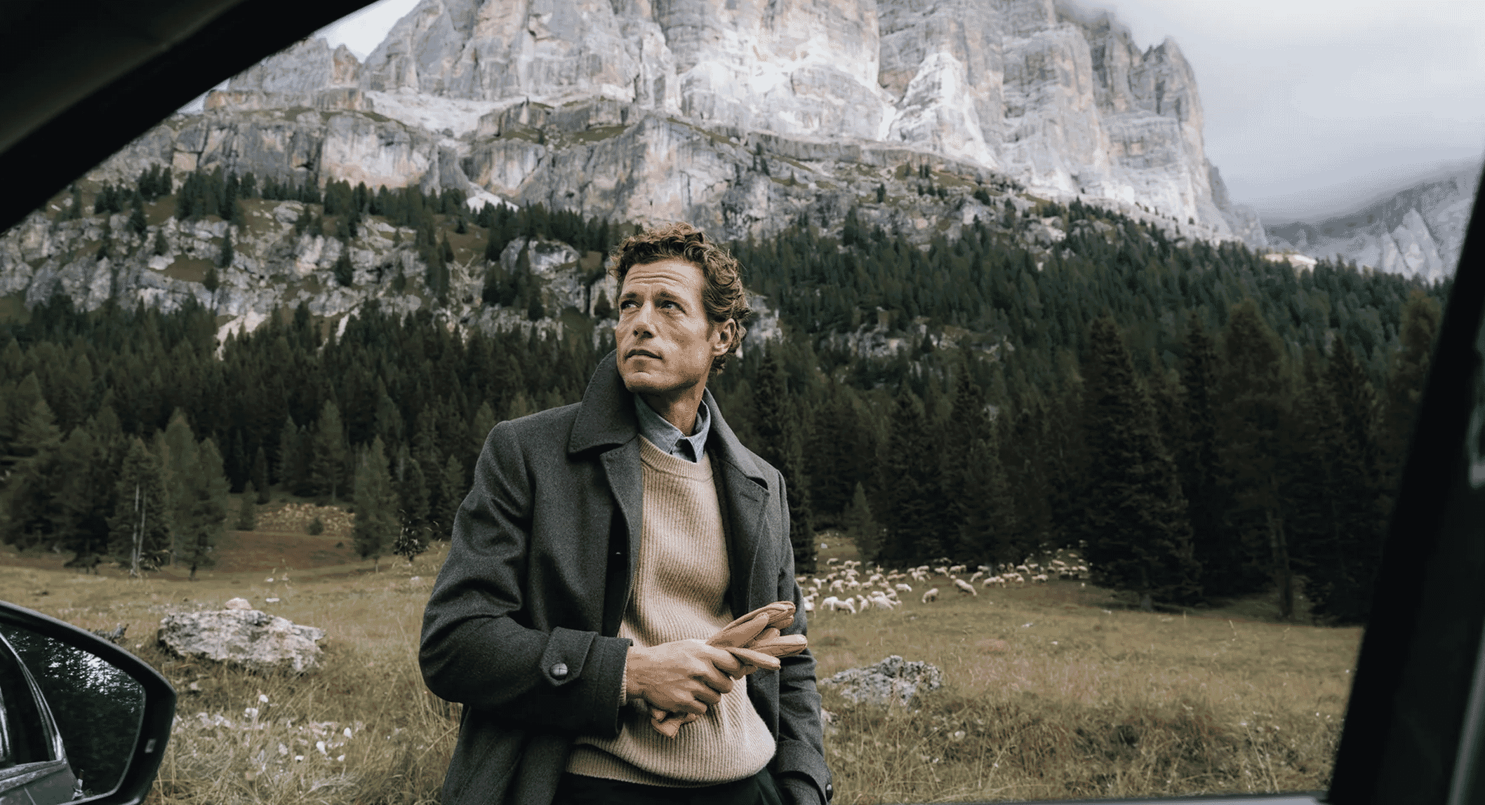 A man in a grey wool coat and beige sweater standing by a car with mountains in the background.