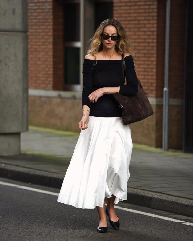 A woman walking in an off-the-shoulder black top and a white midi skirt with a suede tote.