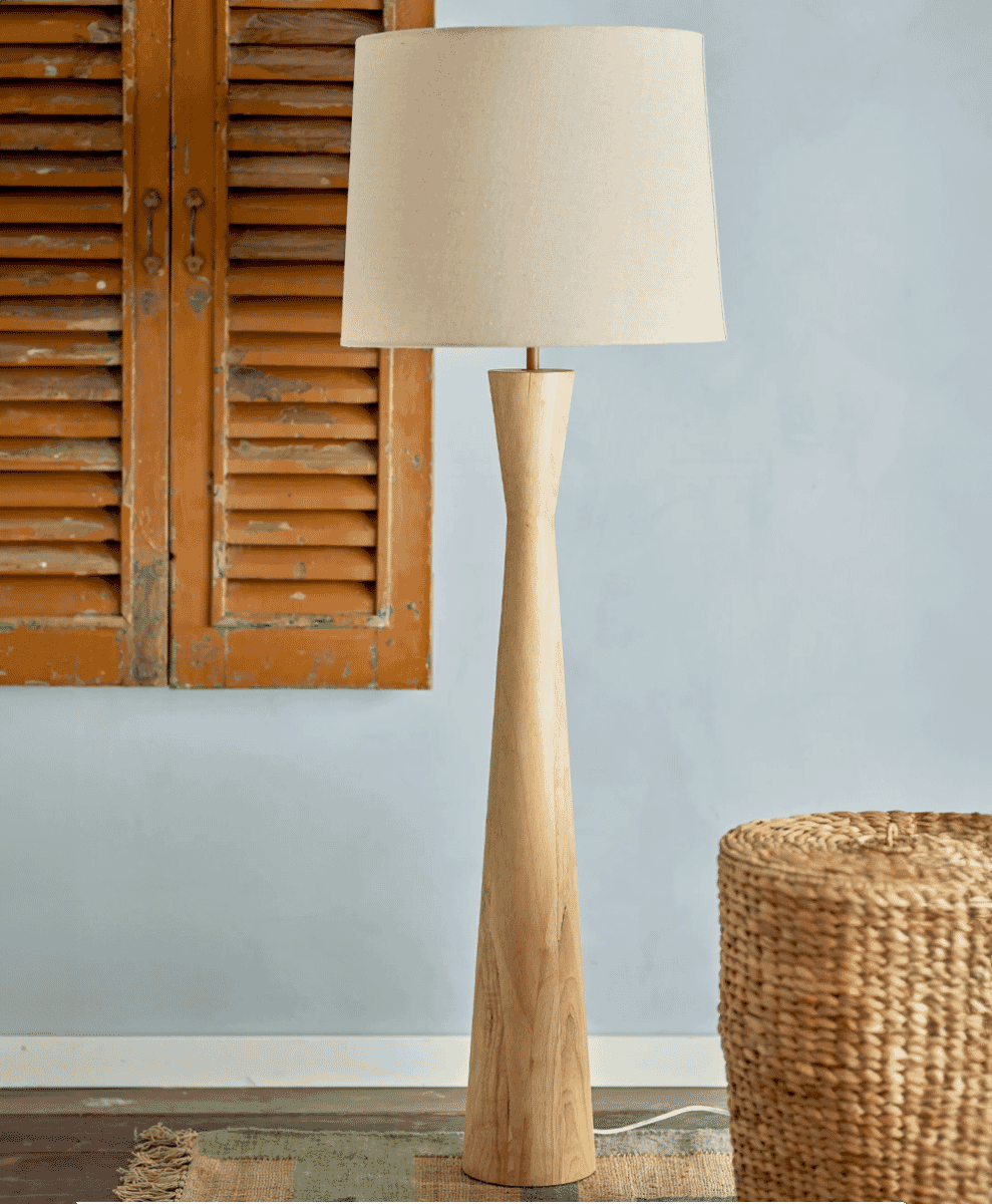 A modern wooden floor lamp with a beige fabric shade in a room with light blue walls and rustic orange shutters.