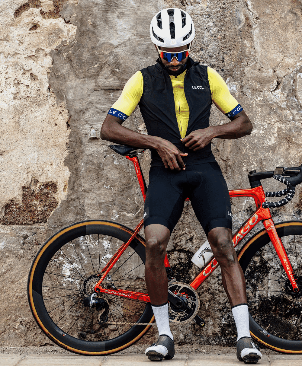 A cyclist in a yellow and black Le Col kit posing with a red road bike against a stone wall.