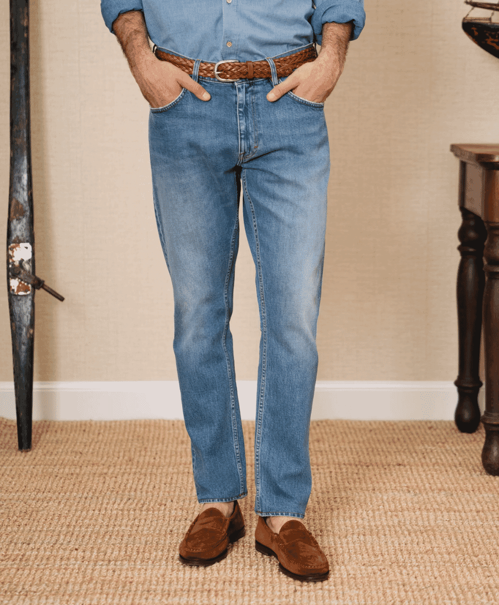 Close-up of a man wearing light-wash blue jeans with brown suede loafers and a braided belt.