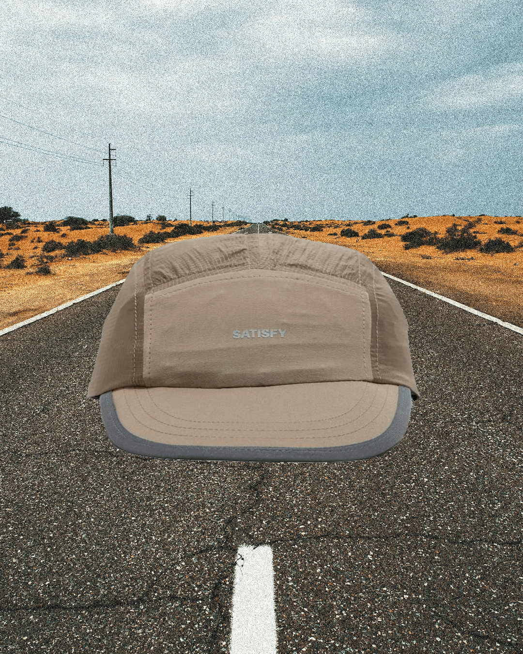 A beige Satisfy five-panel cap centered on an asphalt road in a desert.