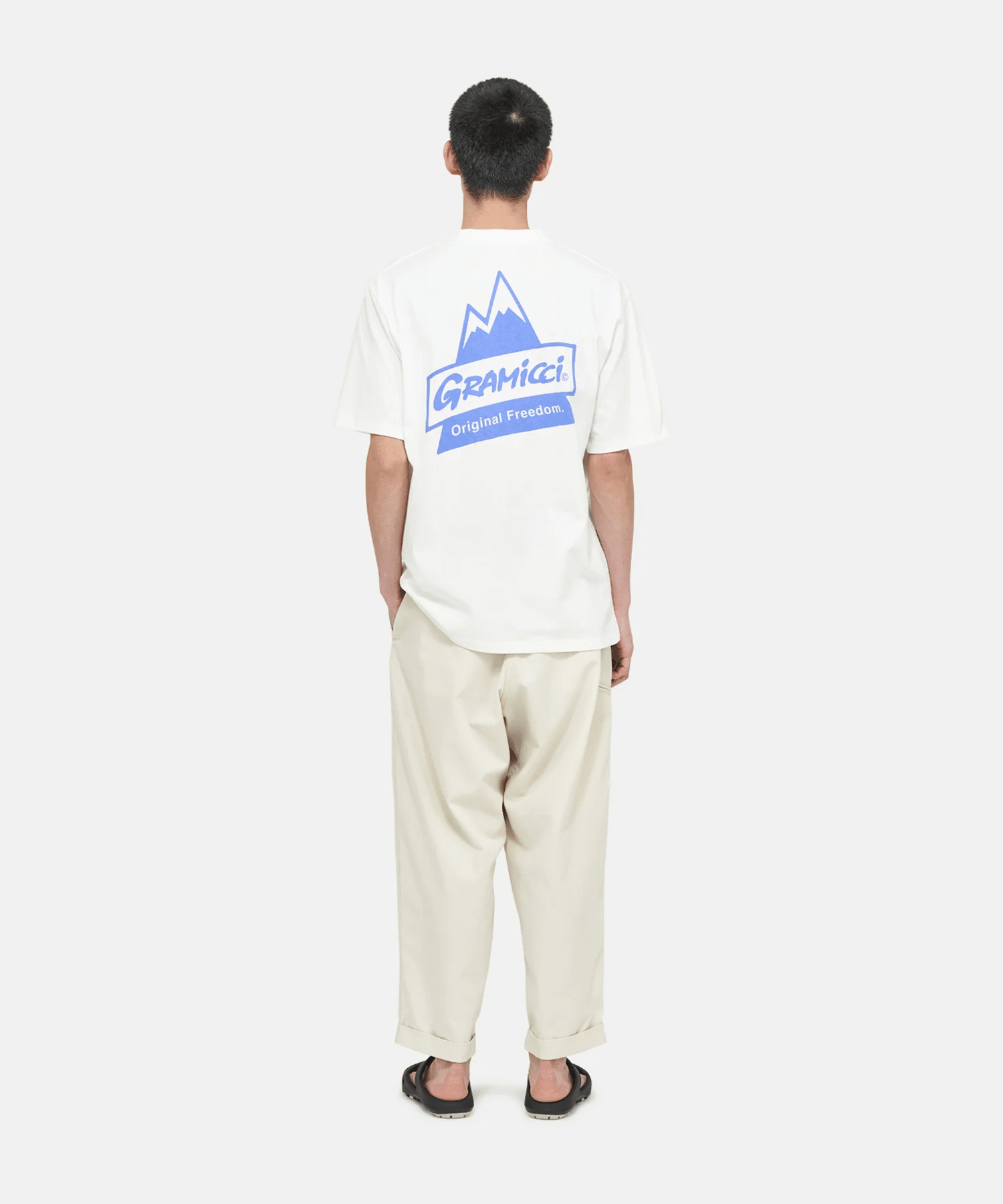 A man viewed from behind wearing a white oversized Gramicci T-shirt with a blue mountain logo.