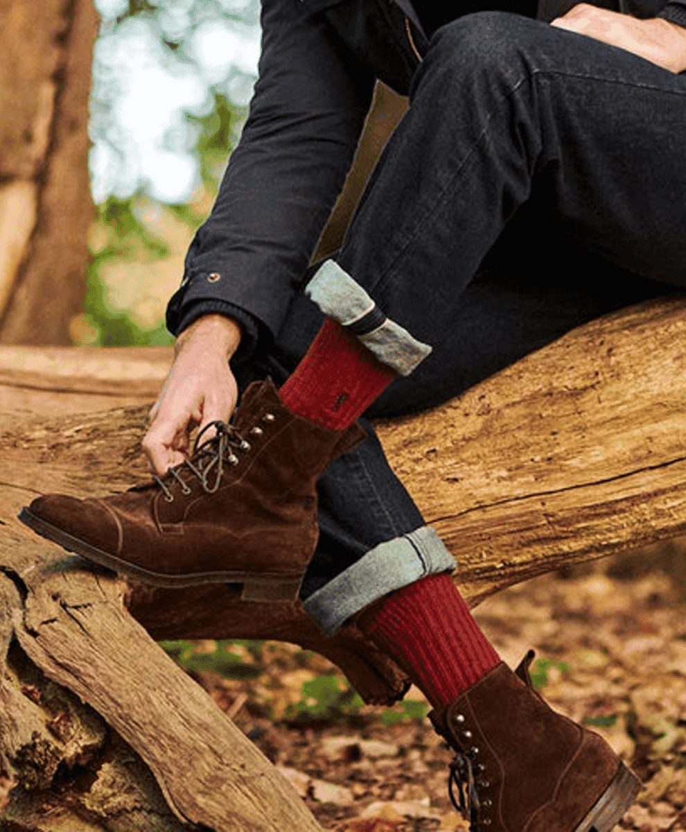 A man in a forest adjusting his brown suede boots, showing red ribbed socks.
