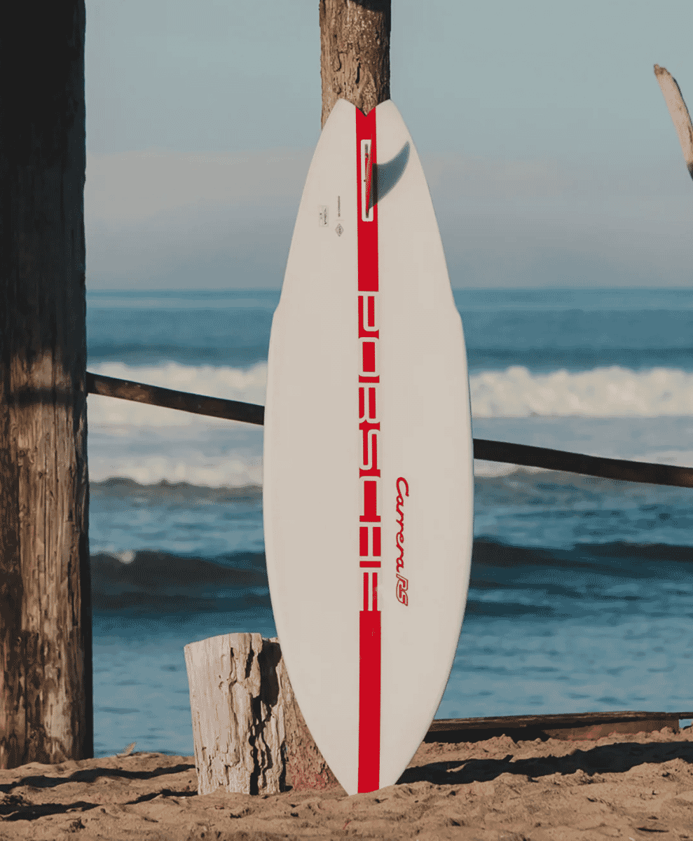 A white surfboard with red accents standing upright on a beach with ocean waves in the background.