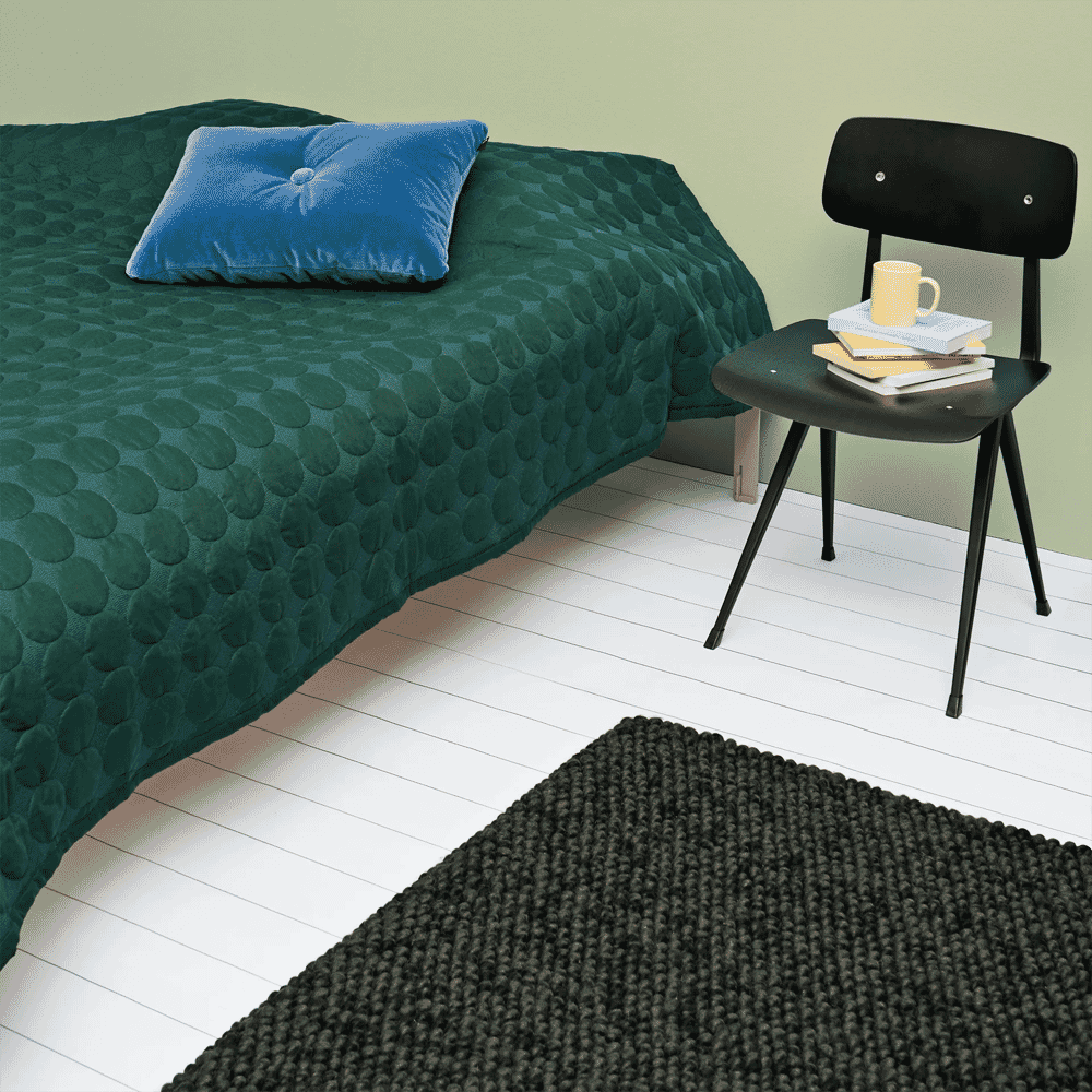 Modern bedroom featuring a green textured quilt, blue velvet pillow, and a dark knitted rug on white wooden floors.