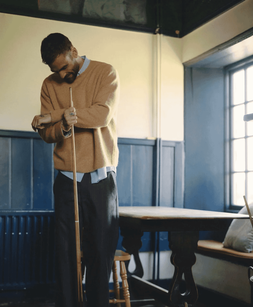 A man in a beige knit sweater over a striped shirt holding a pool cue stick in a rustic pub.