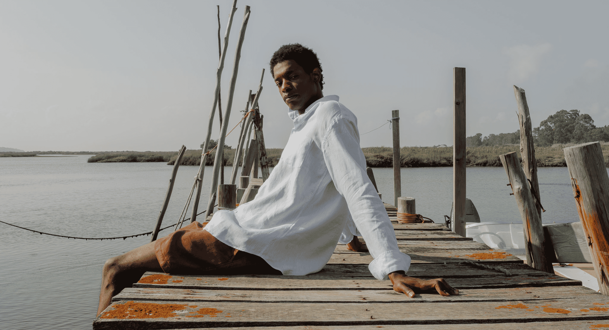 A man in a white linen shirt and brown shorts sits relaxed on a wooden dock by the water.