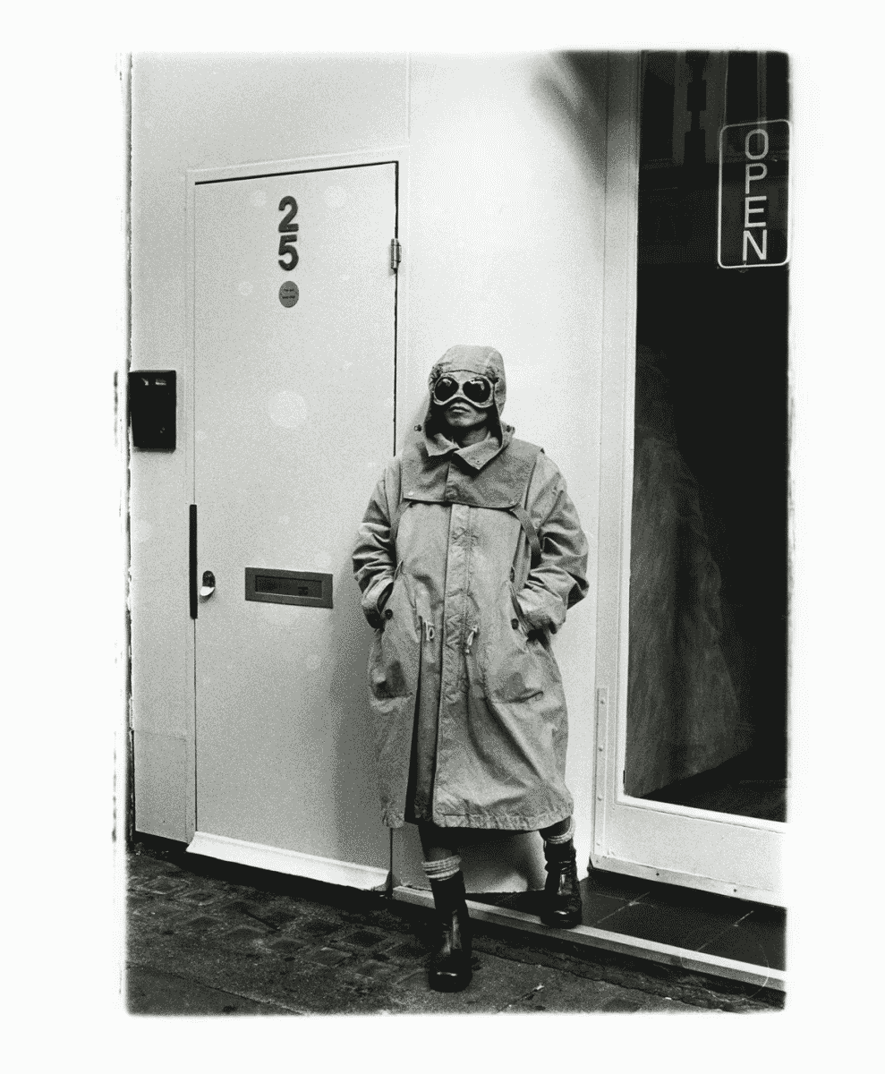 A person in a hooded trench coat and goggles standing near an urban shop entrance.