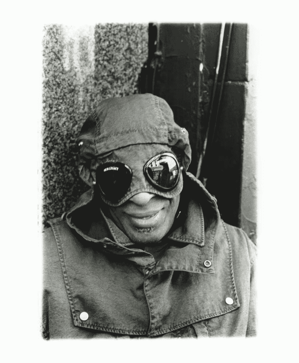 A smiling man wearing a hooded jacket with oversized reflective goggles built into the hood.