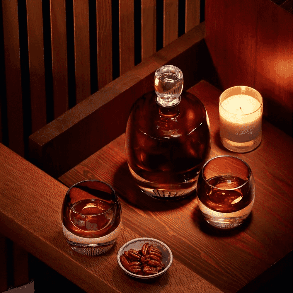 A cozy still life featuring a whiskey decanter, glasses, and a candle on a wooden table.