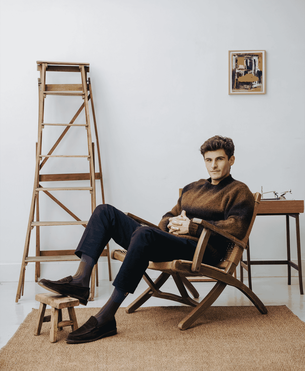 A man in a brown sweater and dark trousers sitting in a minimalist studio setting, showcasing a relaxed tailored look.