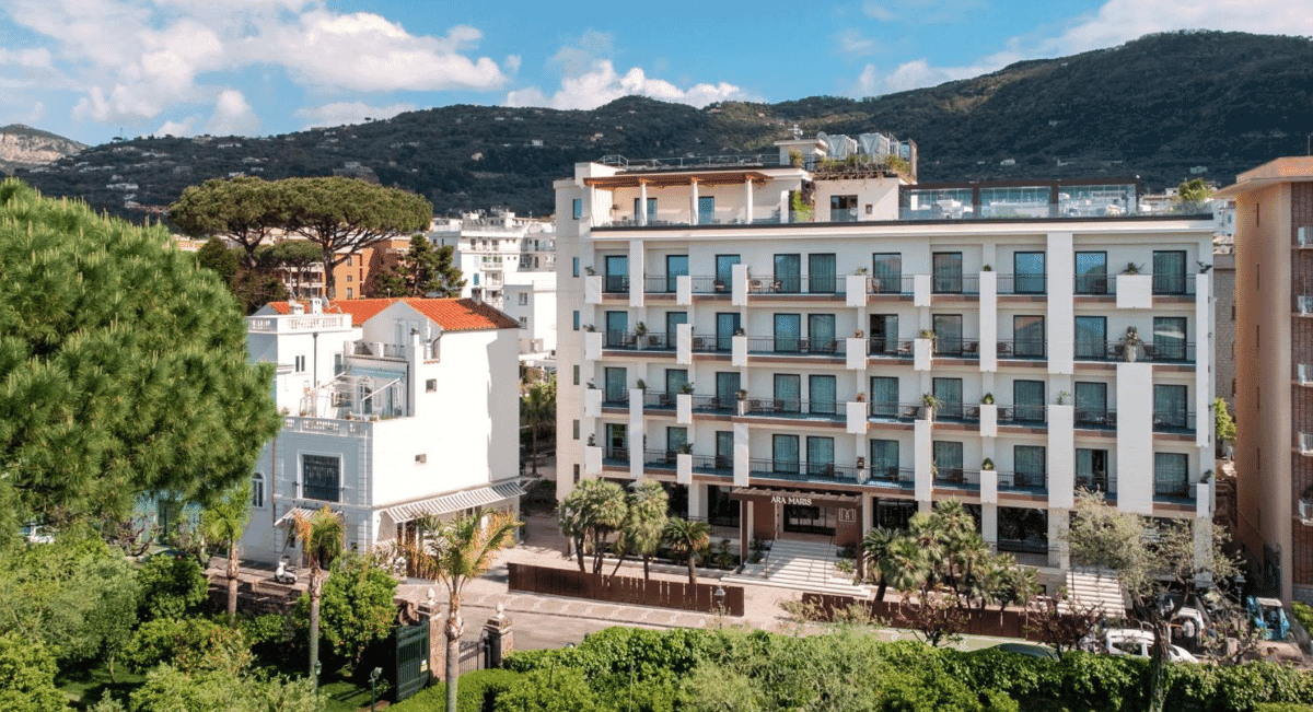 Aerial view of a modern white hotel with balconies surrounded by lush green hills and Mediterranean coastline.