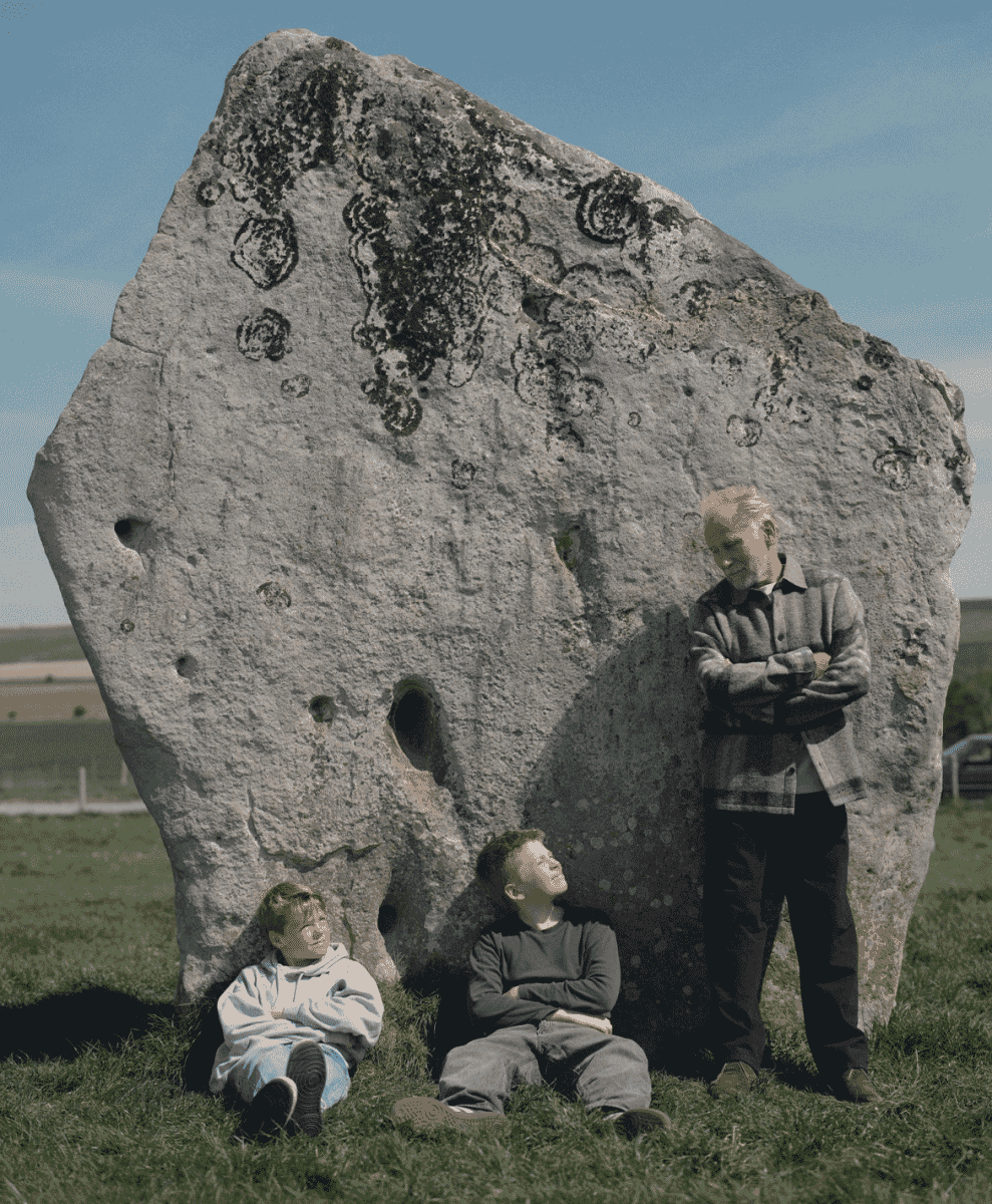 An older man and two boys in casual layered clothing sitting near an ancient stone monument.