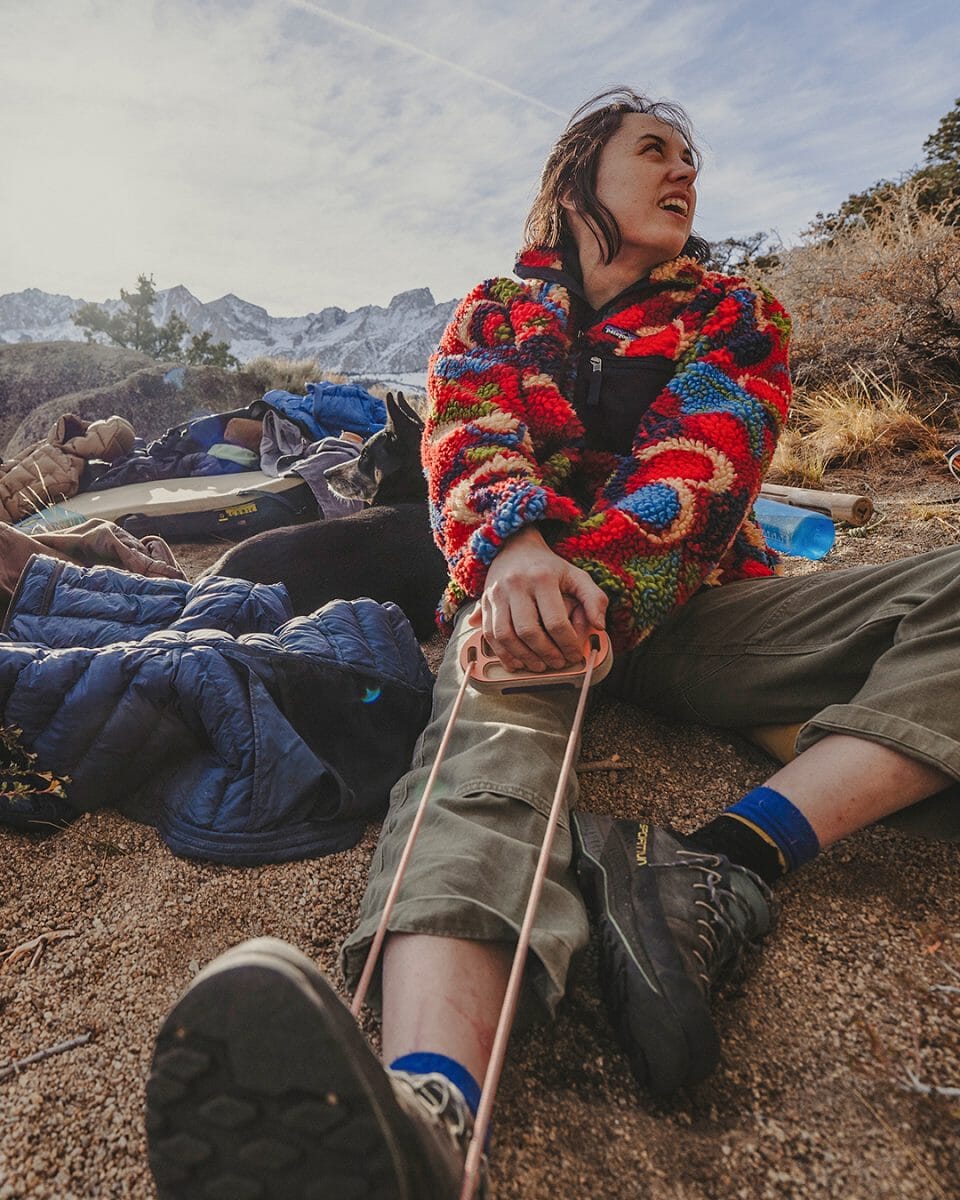 A woman in a vibrant multicolored fleece sitting on rocky ground with camping equipment.