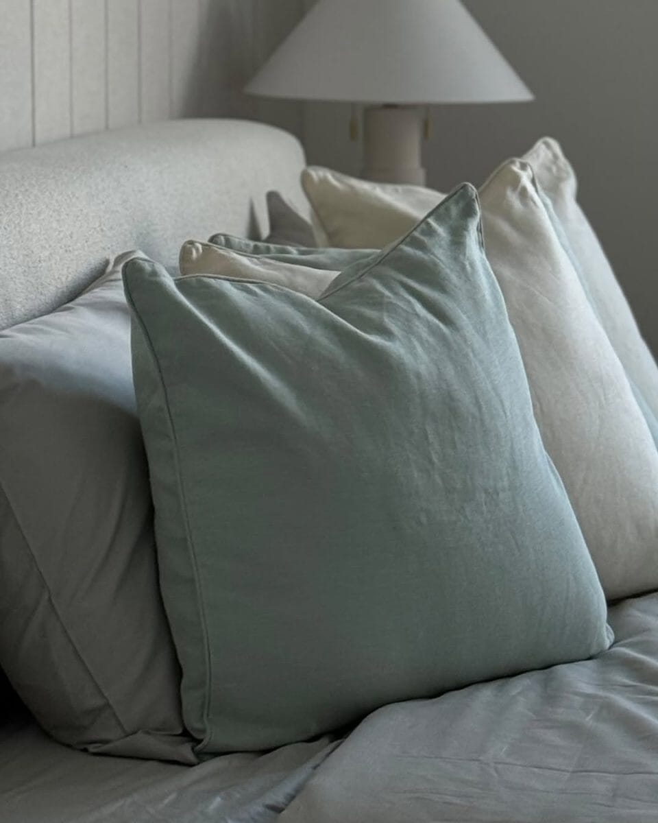 A bed with layered pillows in muted tones of sage green, gray, and cream against a neutral headboard.
