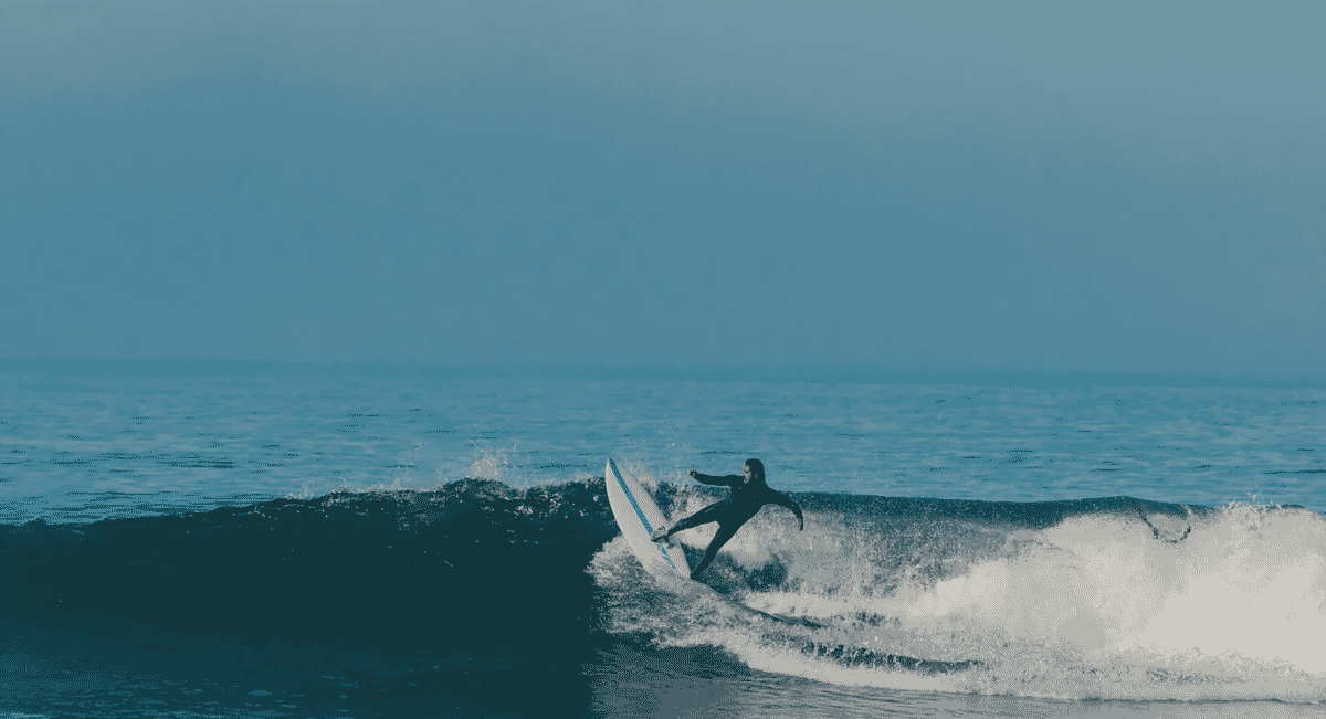 A surfer in a black full-body wetsuit riding a wave on a white surfboard under overcast skies.