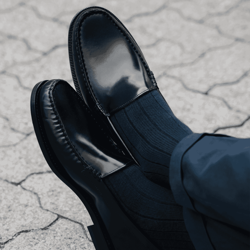 Close-up of glossy black loafers and navy trousers on a stone pavement.